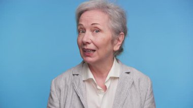 Dost canlısı yaşlı bir kadın, video görüşmesi sırasında kameraya bakarken mavi bir arka planda gülümsüyor..