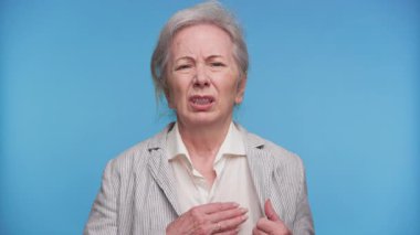 Endişeli yaşlı kadın gözlerini kapatıyor ve göğsünü tutuyor. Muhtemelen mavi arka planda acı çekiyor ya da nefes darlığı çekiyor..