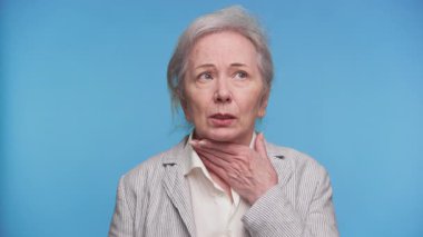 Yaşlı kadın boğazına acı dolu bir ifadeyle dokunuyor, muhtemelen boğazı ağrıyor ya da yutkunmakta zorlanıyor, mavi arka planda..
