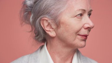 Kameraya gülümseyen yaşlı bir kadının sıcak ve dostane yakın çekimi, yumuşak pembe bir arka planda..