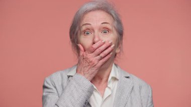 Yaşlı kadın ağzını şoka sokuyor, gözleri sonuna kadar açık, şaşkınlığını ya da inançsızlığını ifade ediyor, pembe bir arka plana karşı duruyor..