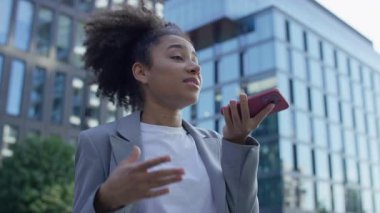 Modern ofis binalarıyla çevrili açık havada sesli bir mesaj kaydederken akıllı telefonu ağzının yanında tutan kendine güvenen bir kadın..