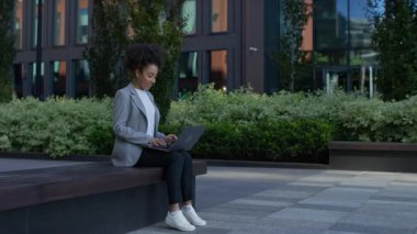 Profesyonel bir kadın, modern bir ofis binasının dışındaki bankta oturuyor. Bilgisayarıyla uzaktan çalışırken odaklanmış ve gülümsüyor..