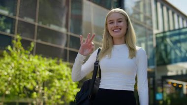 Sırt çantalı genç bir kadın modern binanın önünde duruyor, gülümsüyor ve sıradan beyaz bir bluz giyerken el hareketi yapıyor..