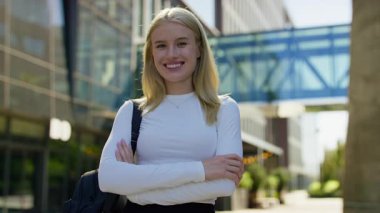 Sırt çantalı gülümseyen genç bir kadın modern bir kampüs binasının önünde kollarını kavuşturup, güpegündüz poz veriyor..