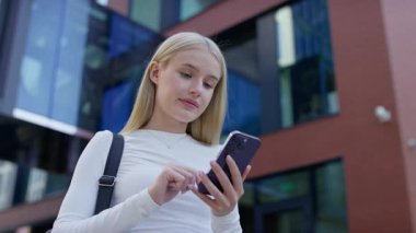 Sırt çantalı öğrenci modern bir binanın önünde duruyor, akıllı telefonunu tutarken ve kaydırırken çevrimiçi içeriği tarıyor.