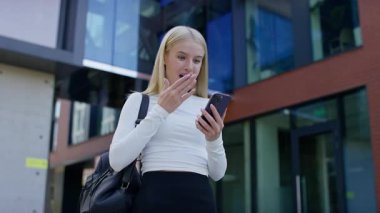 Dışarıdaki gülümseyen öğrenci, akıllı telefonundan iyi haberler okurken sevinçli bir kutlama için yumruğunu kaldırırken heyecanlı görünüyor..