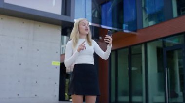 Sarı saçlı bir öğrenci video konuşmasında akıllı telefonunu kullanarak modern bir kampüs binasının önünde dikiliyor..