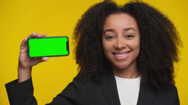 Kendine güvenen, spor ceketli, gülümseyen ve elinde parlak sarı arkaplan ile yeşil ekranlı akıllı bir telefon tutan genç bir kadın..