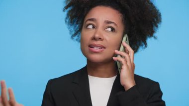 Siyah ceketli genç bir kadın akıllı telefonu kulağına tutuyor, konuşurken gülümsüyor, mavi arka planda dikiliyor..