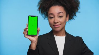 Blazer giyen genç bir kadın yeşil ekranlı akıllı telefonu tutarken mavi arka plana karşı onaylanmamış bir jest yapıyor..