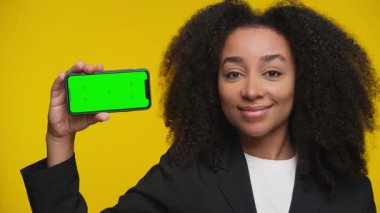 Ceketli gülümseyen genç kadın yeşil ekranlı akıllı telefonu tutarken el hareketi yapıyor, parlak sarı arka planda duruyor..