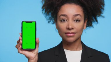 Ceketli, kendine güvenen genç bir kadın kameraya doğru açık yeşil ekranlı akıllı telefonu tutuyor. Parlak mavi arkaplanda duruyor. Model için mükemmel..