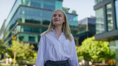 Beyaz gömlekli ve gri pantolonlu gülümseyen genç bir kadın arka planda cam ofis binaları ve yeşillik olan modern bir şehirde güvenli bir şekilde yürüyor..