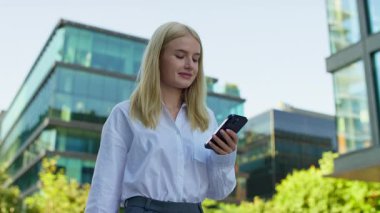 Beyaz tişörtlü sarışın kadın dışarıda akıllı telefon kullanıyor, modern cam ofis binalarının yanında yürüyor, online içerikleri araştırmaya odaklanmış..