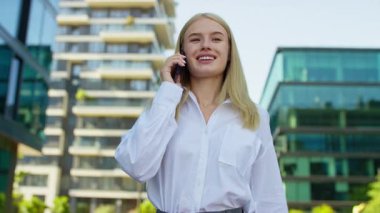 Beyaz tişörtlü, kendine güvenen, akıllı telefonlu, gülümseyen, modern şehir ofisi ve apartmanlarının yanında duran sarışın kadın..