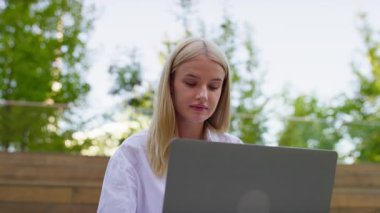 Genç profesyonel dizüstü bilgisayarla ahşap basamaklarda oturuyor, yeşil ağaçlar ve doğal ışıklarla çevrili bir açık hava ortamında çalışıyor..