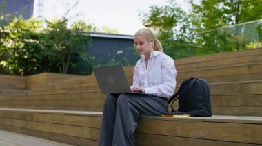 Genç bir kadın dizüstü bilgisayarla tahta basamaklarda oturuyor, kulaklık takıyor ve çalışırken ya da sırt çantasıyla çalışırken gülümsüyor..