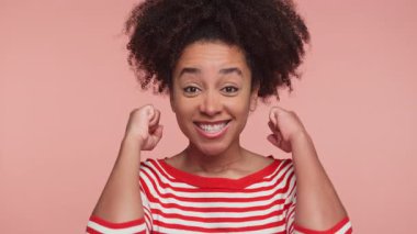 Çizgili süveter giymiş gülümseyen genç bir kadın, pembe arka planda heyecan ve mutluluk gösteren bir kutlama için yumruklarını kaldırıyor..