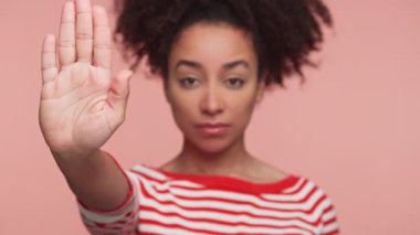 Kırmızı beyaz çizgili süveter giyen ciddi genç bir kadın avucunu ileri doğru kaldırıyor. Pembe bir arka plana karşı sert bir yüz ifadesiyle açık bir dur ya da dur hareketi yapıyor..