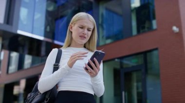 Modern ofis binasının önünde duran genç profesyonel kadın, endişeli bir şekilde kafasını tutarken akıllı telefondan stresli ya da kötü haberler okuyor..