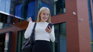 Sırt çantalı, gülümseyen ve kutlama yapan genç bir kadın modern bir binanın dışında akıllı telefondan olumlu haberler ya da mesajlar okuyor..