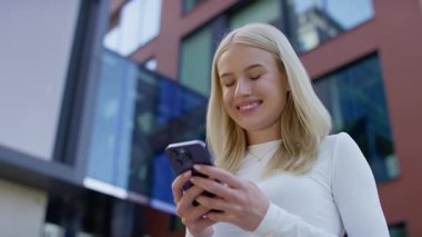 Modern binanın önünde duran genç bir kadın, gülümseyerek akıllı telefonuna rahat ve rahat bir şekilde bir mesaj yazıyor..