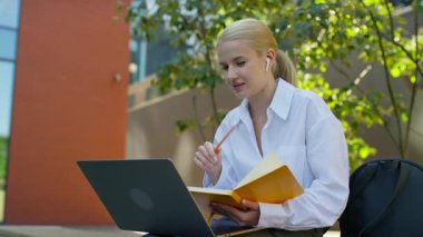 Bilgisayarlı ve defterli genç bir kadın, kulaklık takıyor, dışarıda online bir derse katılırken konuşuyor ve notlar yazıyor..