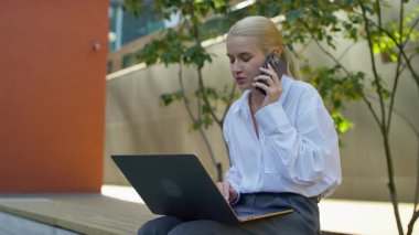 Dışarıda dizüstü bilgisayarla oturan, çalışırken ya da okurken akıllı telefonla konuşan, modern bir ortamda görevleri yöneten genç bir kadın..