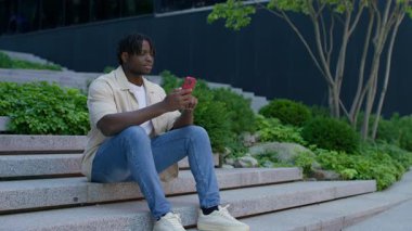 Neşeli genç adam dışarıda merdivenlerde oturuyor, yumruğunu kaldırıyor ve neşeyle gülüyor, akıllı telefonu tutuyor, başarıyı veya başarıyı kutluyor..