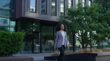 Şık giyinen profesyonel genç kadın şehir ortamındaki modern cam ofis binasının yanında güvenle yürüyor..