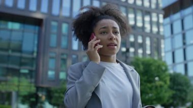 Profesyonel kıyafetli, modern şehir ortamında akıllı telefondan konuşan, arka planda ofis binaları olan odaklanmış bir kadın..