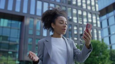 Spor ceketli neşeli genç bir kadın modern cam ofis binasının önünde video görüşmesinin keyfini çıkarıyor..
