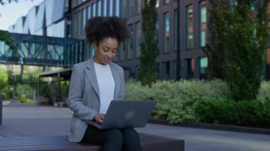 Spor ceketli bir iş kadını, modern ofis binalarının dışındaki bankta otururken dizüstü bilgisayar kullanıyor, uzaktan çalışmayı ve üretkenliği temsil ediyor..