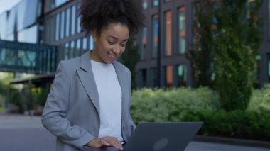 Spor ceketli neşeli bir iş kadını modern bir ofis binasının önünde çalışırken ya da çalışırken laptop ekranına bakıyor..