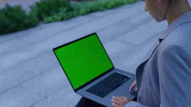 Spor ceketli profesyonel bir kadının, yeşil ekranlı bir dizüstü bilgisayar kullanarak, modern bir şehir ortamında, açık hava merdivenlerinde oturduğu yakın plan..