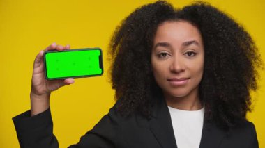 Kendine güvenen spor ceketli genç kadın canlı sarı arka plana karşı yeşil ekran ekranlı akıllı telefonu tutuyor, uygulama modellemeleri ve sunumlar için kullanışlı.