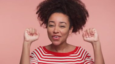 Kırmızı çizgili tişörtlü neşeli genç kadın enerjik bir ifadeyle dans ediyor. Pembe arka planda coşku ve pozitiflik yayıyor..