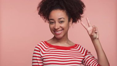 Kırmızı çizgili tişörtlü neşeli genç kadın pembe arka planda parmakları olan barış tabelasını tutarken ışıl ışıl gülümsüyor..