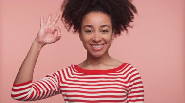 Kırmızı çizgili tişörtlü neşeli genç bayan pembe arka planda normal bir el işareti yaparken kendine güvenerek gülümsüyor..