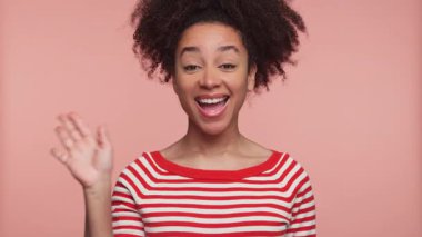 Kırmızı çizgili gömlekli neşeli genç kadın pembe arka planda gülümserken el sallıyor..