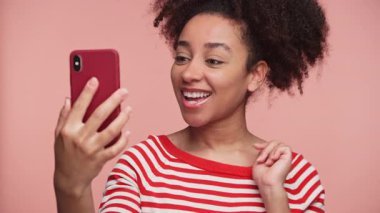 Kırmızı çizgili tişörtlü mutlu genç bir kadın pembe arka planla konuşurken kırmızı çantalı akıllı telefonu tutuyor..