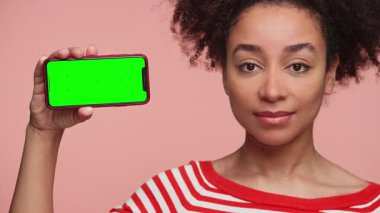 Kırmızı çizgili gömlekli kendine güvenen genç bir kadın, pembe arka planda yeşil ekranlı akıllı telefonu sunar, uygulama veya tasarım modeli için idealdir..