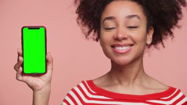 Kırmızı çizgili tişörtlü mutlu genç kadın pembe arka planda yeşil ekranlı akıllı telefonu gösterirken onaylıyor..