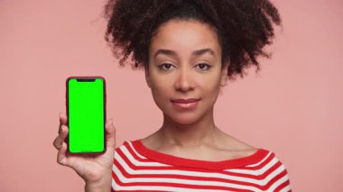 Elinde boş yeşil ekranlı akıllı telefon tutan genç bir kadının portresi, pembe arkaplana karşı model veya uygulama sunumuna hazır..