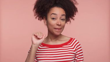 Çizgili tişörtlü, mutlu genç bir kadın gülümseyip pembe bir arka plana veda ederken el sallıyor..