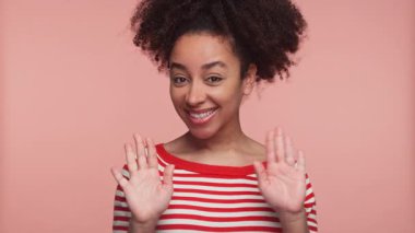 Çizgili tişörtlü neşeli genç bayan pembe bir arka plana karşı iki elini sallıyor ve gülümsüyor..