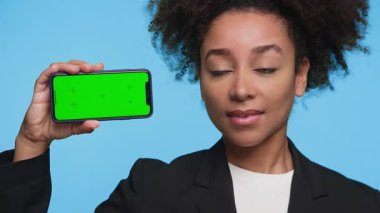 Siyah ceketli genç bir kadın elinde yeşil ekranlı akıllı telefon tutarken mavi arka plana karşı kafasını sallıyor..