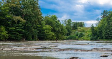 Kırsal alan. Dramatik bir gökyüzünün altında sığ bir nehrin yakınındaki ağaçlar.