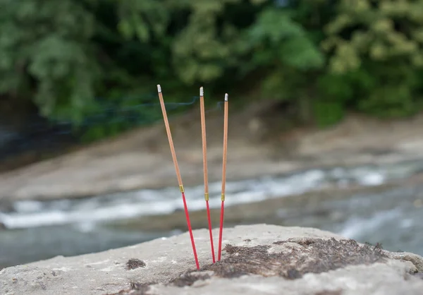 Three Buddhist burning incense sticks in the stones on the river Bank. Selective focus.